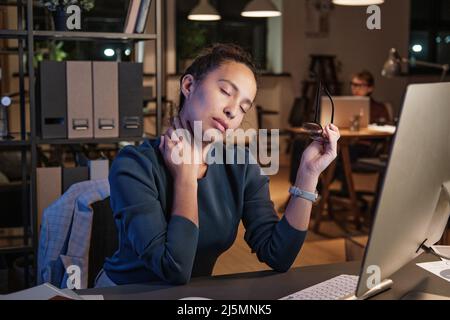 Tired young mixed race woman with closed eyes sitting with glasses at table and touching neck while feeling its pain, working late concept Stock Photo