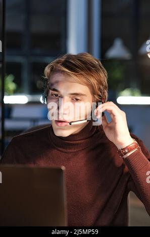 Handsome young call center operator in brown sweater adjusting headset microphone while working at night Stock Photo