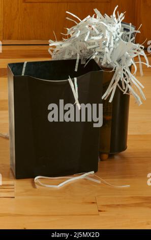 Paper waste shredder basket and wastebasket overflowing with shredded paper strips in home office setting. This closeup shows a wood floor background. Stock Photo
