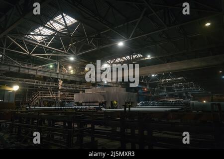 Large girder or beam crane transports iron pipes inside large dark workshop in metallurgical factory. Stock Photo