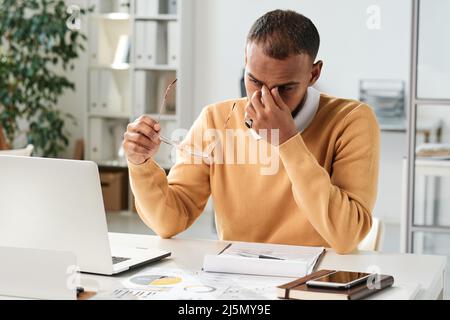 Tired young middle eastern businessman holding eyeglasses and rubbing bridge of nose while overworking in office Stock Photo