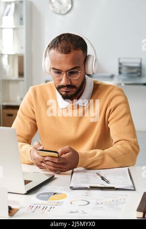Serious young middle eastern businessman in headphones and eyeglasses sitting at table with papers and checking smartphone Stock Photo