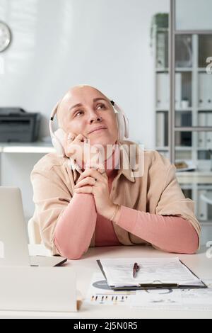 Dreamy young businesswoman in wireless headphones sitting at table with laptop and looking up in modern office Stock Photo