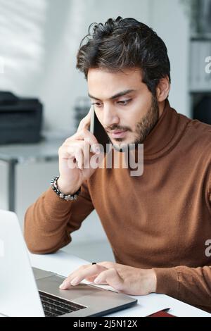 Serious modern businessman in brown turtleneck sitting at table and using laptop while having phone conversation Stock Photo
