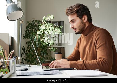 Confident young middle eastern manager in sweater typing on laptop while working in office Stock Photo