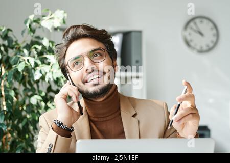 Positive handsome young middle eastern entrepreneur with beard holding pen while talking on phone in office Stock Photo