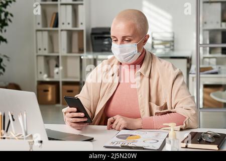 Concentrated young Caucasian bald woman in mask sitting with smartphone at table while working with business papers in office Stock Photo