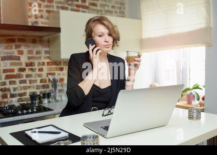 A young blond woman works remotely doing calls in her kitchen at home. Stock Photo