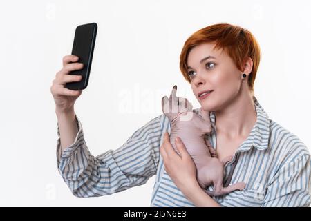 Redhead young woman hugging Sphynx Cat, taking selfie photo on cellphone, looking at telephone camera. Studio shot on white background Stock Photo