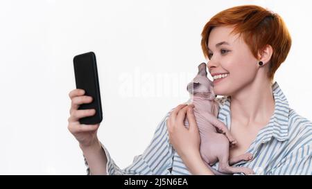 Smiling redhead young woman hugging Sphynx Cat, takes selfie photo on smartphone, looking at mobile phone camera. Beautiful hipster Stock Photo