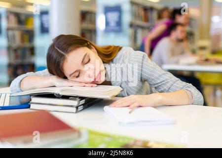 Tired young woman lying on stack of books in library Stock Photo