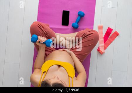 Pregnant woman training at home with online fitness class on phone app doing bicep curl workout. Top view of Asian girl working out during pregnancy Stock Photo