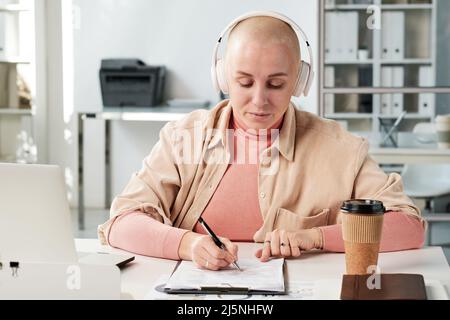 Creative web designer in wireless headphones writing down ideas into paper in office Stock Photo