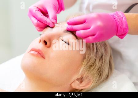 Mesotherapy and rejuvenation. Portrait of an adult blonde woman at a cosmetologist's appointment. A beautician in pink medical gloves applies a needle Stock Photo