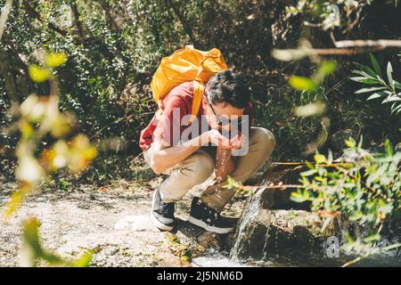 Adult man drinking water from the mountain creek. Tourist wearing casual clothes and sunglasses making a sip of mountain river water from the palms of Stock Photo