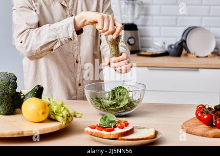 Close-up of young woman adding spices in salad with vegetables, she standing at table in kitchen and preparing food for dinner Stock Photo