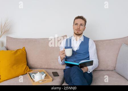 Focused young business man sits on couch with notebook in his hand and reads and analyzes an action plan for development at work in a blue suit. Caucasian businessman in the office on the couch Stock Photo