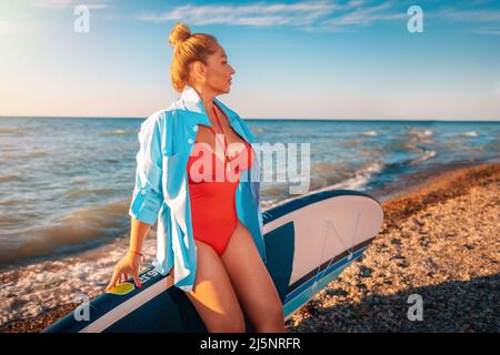 Portrait of pretty plump caucasian woman posing with a sup board. In the background, the sky is the ocean. The concept of sports recreation. Stock Photo