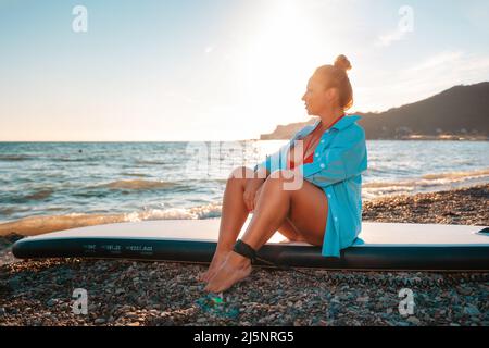 A plump caucasian woman in shirt poses sitting on a sup board. Sunset and ocean in the background. The concept of sports recreation by the sea. Stock Photo