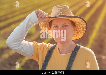 Portrait of female corn farmer in cultivated maize field wearing straw hat and jeans bib overalls and standing among young crop seedlings Stock Photo