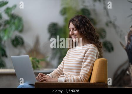 Freelancer young woman sitting in armchair in greenhouse, working on laptop surrounded by houseplant Stock Photo