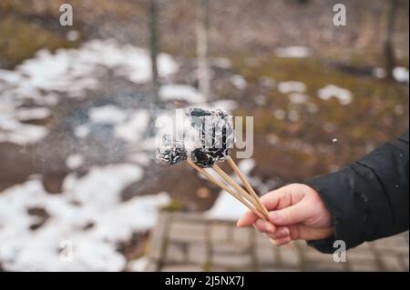 An adult woman's hand holds a three burning paper flowers in front of a winter park. The black charred paper is smoking Stock Photo