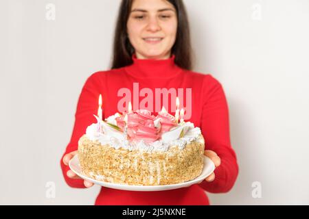 A brunette woman in red clothes holds a large white cake with candles, makes a wish Stock Photo