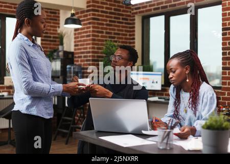African american assistant offering to project manager a cup of coffee while in modern meeting room. Office workers discussing about financial data and management plan of research business. Stock Photo