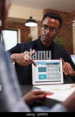 Marketing and research agency group leader pointing out business statistics data on tablet. African american business man informing office colleague about necessity of budget cutting. Stock Photo