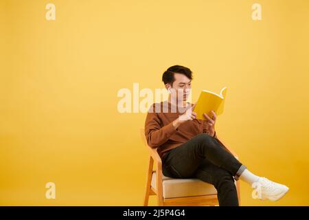 Studio portrait of young Asian man sitting on armchair and reading book Stock Photo
