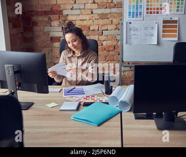 Portrait of cheerful woman, interior designer holding, looking at color samples, sitting at the desk in her office Stock Photo