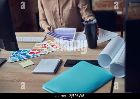 Cropped shot of interior designer working on project, looking at color swatches, holding bottle while sitting at workplace Stock Photo