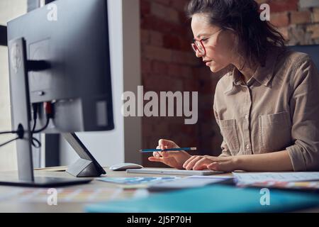 Side view of creative woman, interior architect or designer wearing glasses working on a project, using computer while sitting at the desk in her Stock Photo