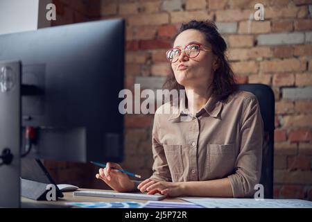Thoughtful female interior architect or designer working on a project, using computer while sitting at the desk in her office Stock Photo