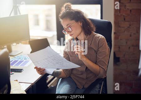 Focused young woman designer holding and studying blueprint paper while working on new interior design project in her office Stock Photo