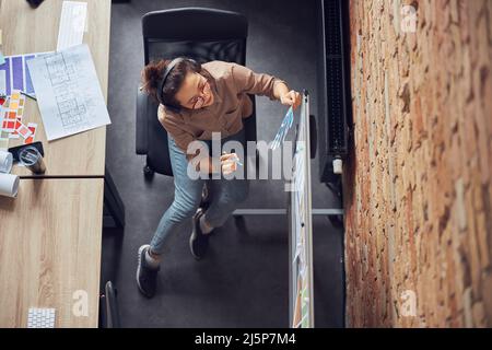 Top view of professional female designer wearing headphones selecting color from samples, looking at blueprint on a board while working on new Stock Photo