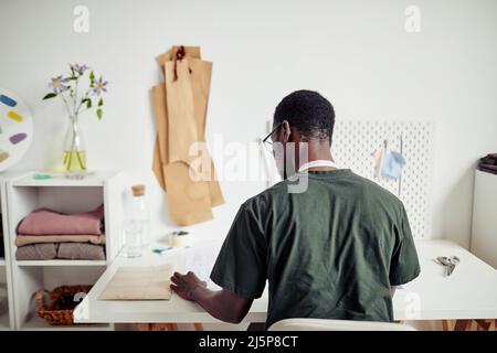 Young tailor sitting at desk in his studio and choosing sewing pattern for his next project Stock Photo