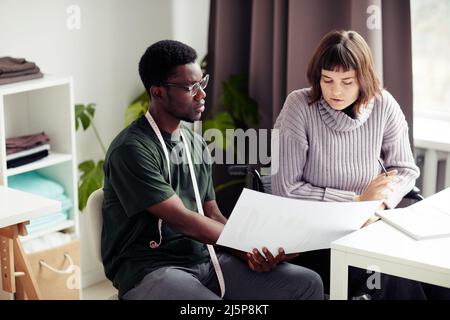 Diverse team of young fashion designers discussing sketches of clothes for new collection at morning briefing Stock Photo