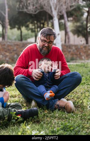Father enjoying playing with his children sitting on the grass outdoors. Stock Photo