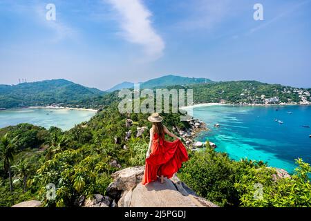 Woman in long red dress and hat standing to pose on high rock at a popular viewpoint on Nang Yuan Island Stock Photo