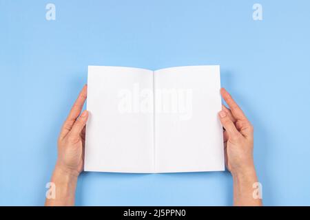 Woman hands holding folded blank paper sheet, empty booklet, double-pages book, magazine on light blue background. Mock up template. Top view Stock Photo
