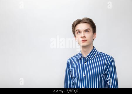 Portrait of thoughtful young man wearing blue-white striped button shirt looking up. Studio shot on gray background Stock Photo