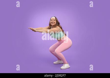 Happy young pretty european plus size female in sportswear squats, isolated on purple background Stock Photo