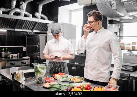 Young chef in uniform checking freshness of vegetables standing near kitchen table with his assistant making notes behind him Stock Photo