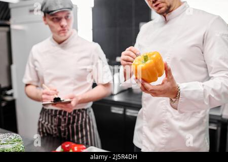 Close-up of chef in uniform holding fresh pepper in his hands and talking to his assistant during cooking Stock Photo
