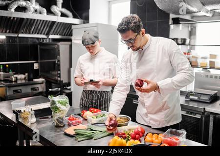 Young chef in uniform choosing fresh vegetables for dish at table with his assistant making notes on paper, they working in commercial kitchen Stock Photo