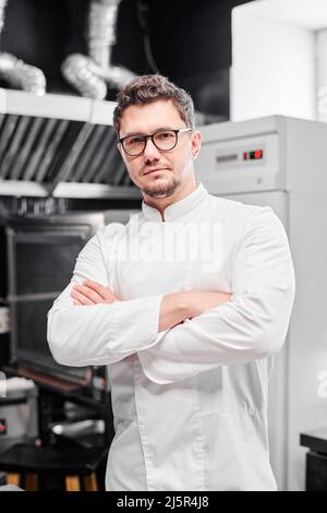 Portrait of young chef in eyeglasses and white uniform standing with his arms crossed in kitchen and looking at camera Stock Photo