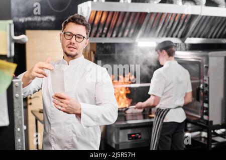 Chef in eyeglasses and white uniform reading order of guest for his assistant who using oven in background Stock Photo