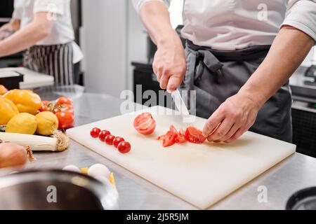 Close-up of young cook in apron using knife to cut fresh tomatoes on cutting board at table Stock Photo