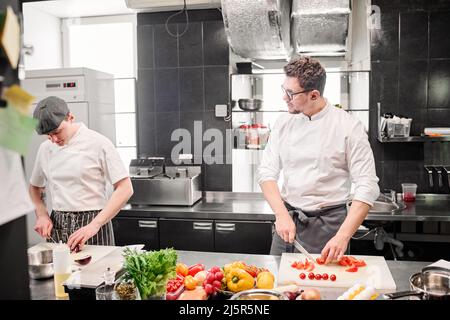 Young chef cutting tomatoes on cutting board and discussing menu with his assistant during their work at table in kitchen Stock Photo